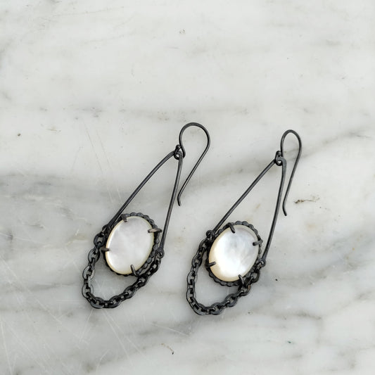 A pair of earrings featuring chains and mother of pearl centers, with black patina on silver metal, displayed on a marble surface.