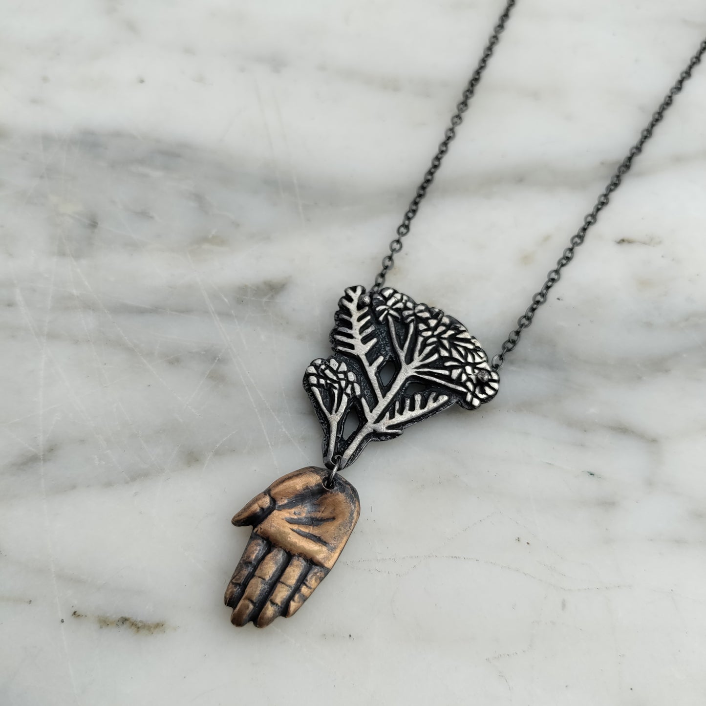Bronze hand and silver yarrow pendant on marble background by Alice Scott.