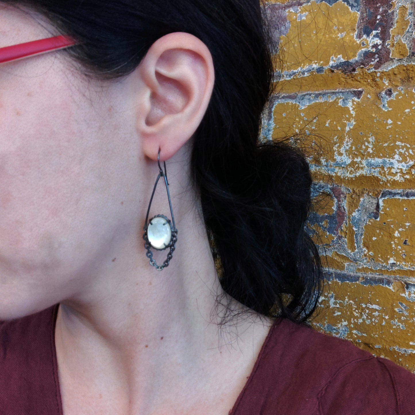 A pair of earrings featuring chains and mother of pearl centers, with black patina on silver metal, displayed on a model ear by Alice Scott