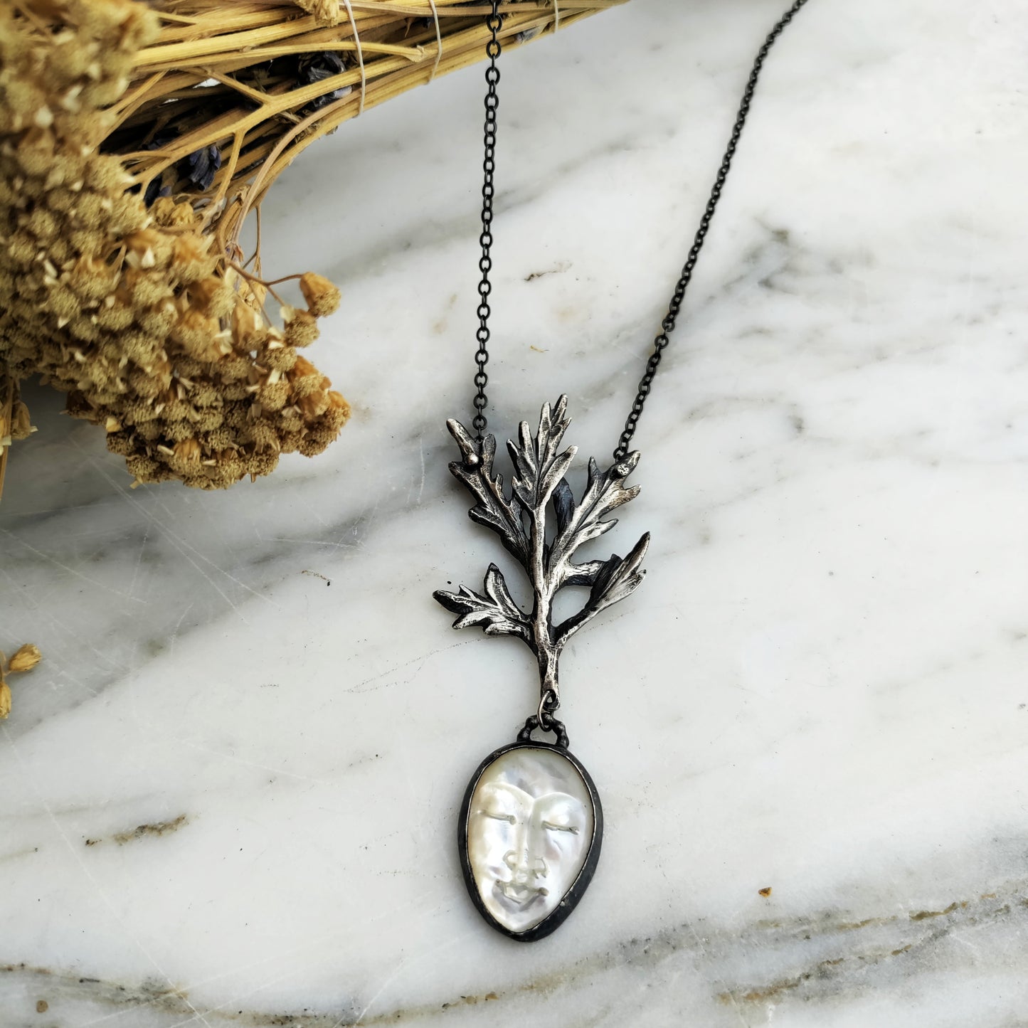 A silver necklace featuring a pearlescent lady mother of pearl pendant, and carved silver mugwort with a black patina chain on a marble background by Alice Scott