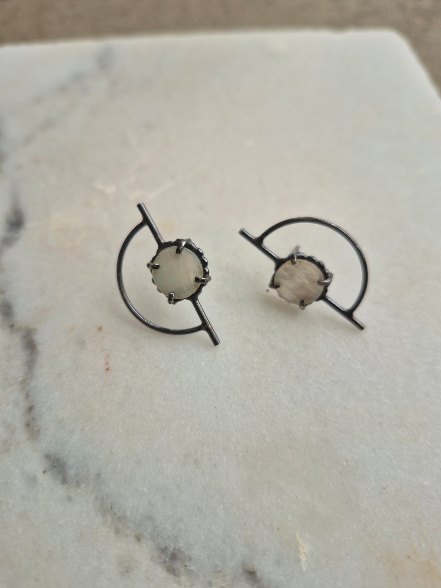 A pair of earrings featuring circular mother of pearl centers with orbiting black post settings, displayed on a marble surface by Alice Scott
