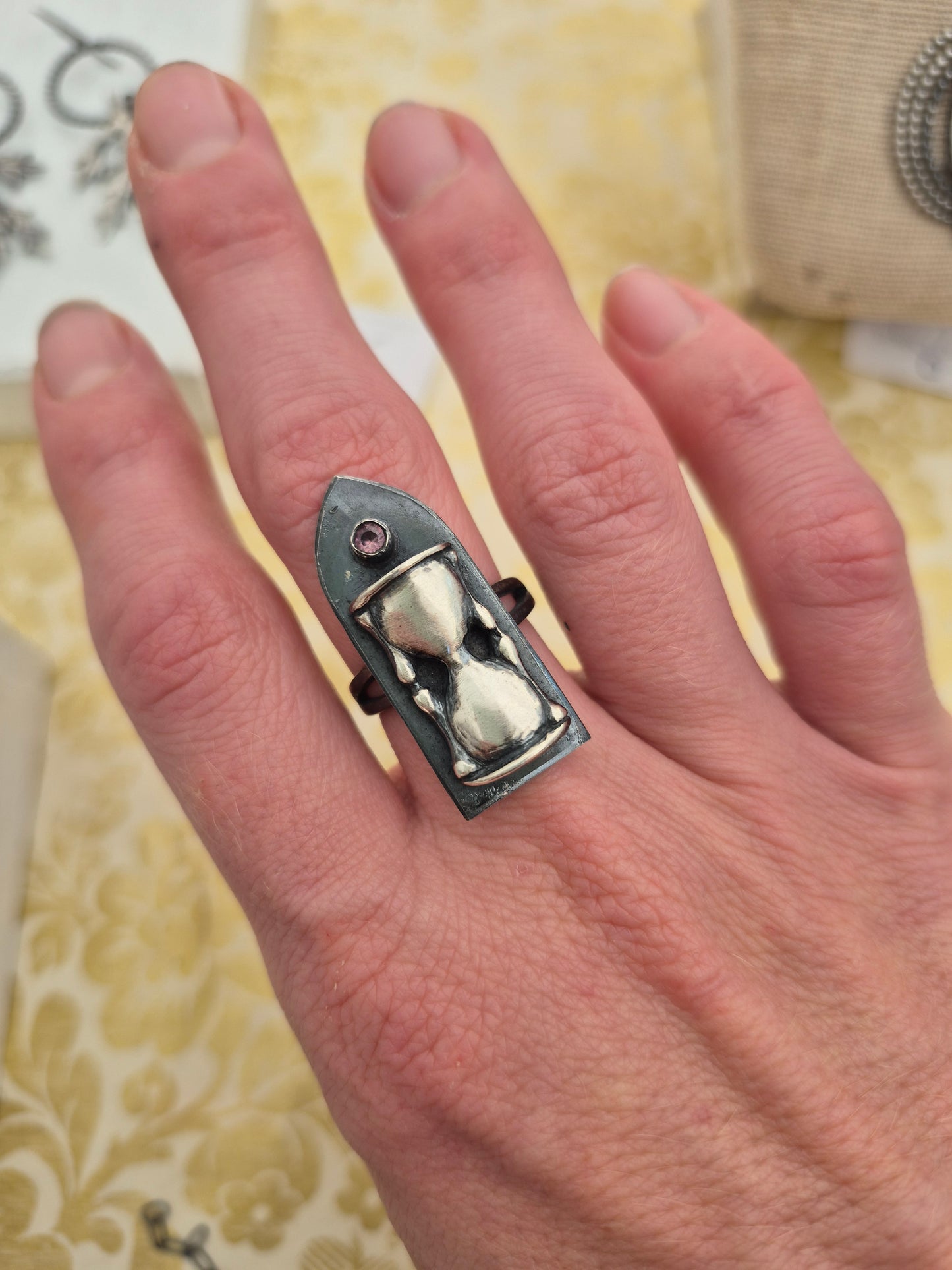 Hand wearing a unique ring with a hourglass design with a pink sapphire on a patterned background, by Alice Scott