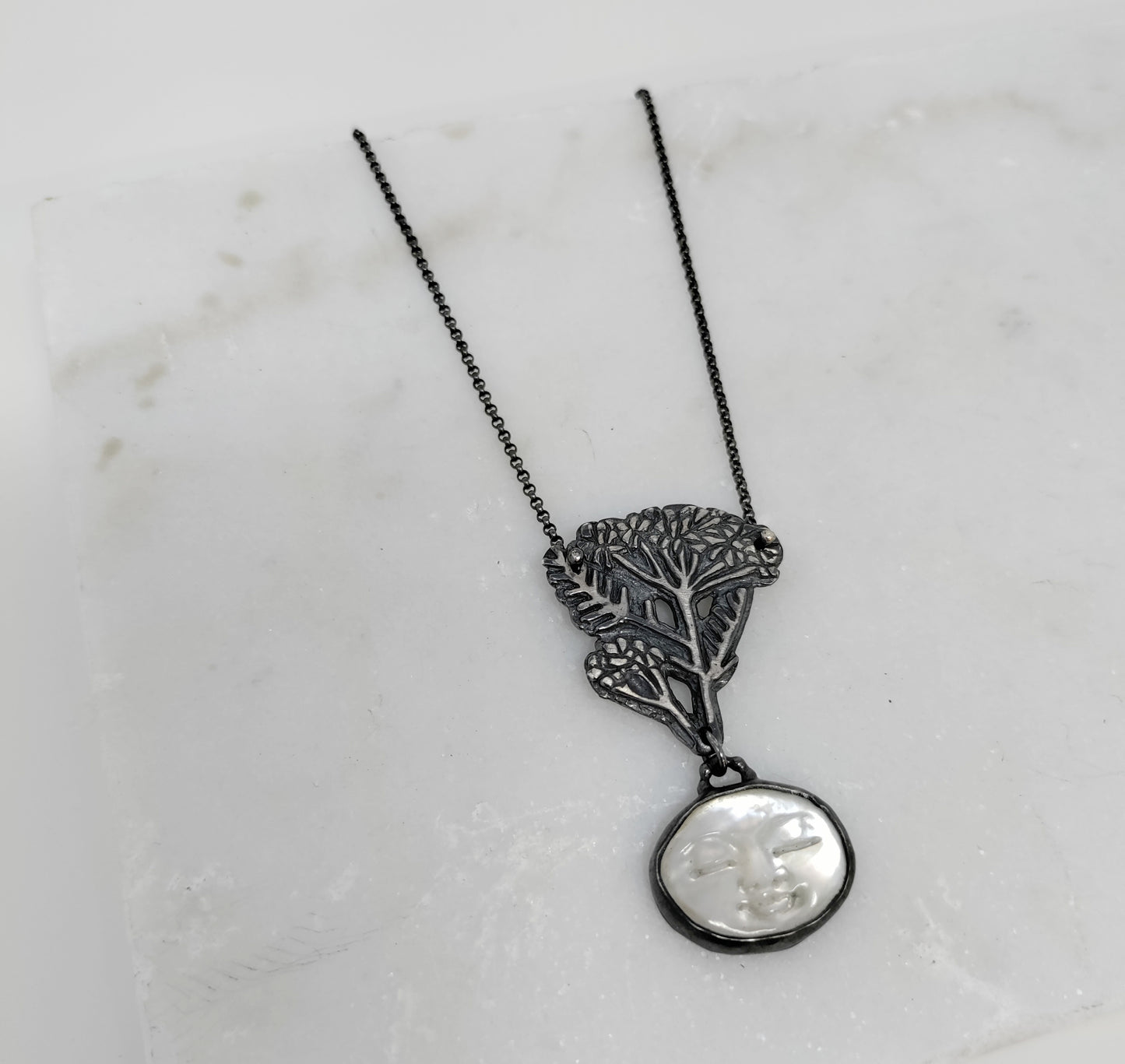 silver necklace with carved mother of pearl moon face and blacked silver yarrow design on a marble background, by Alice Scott