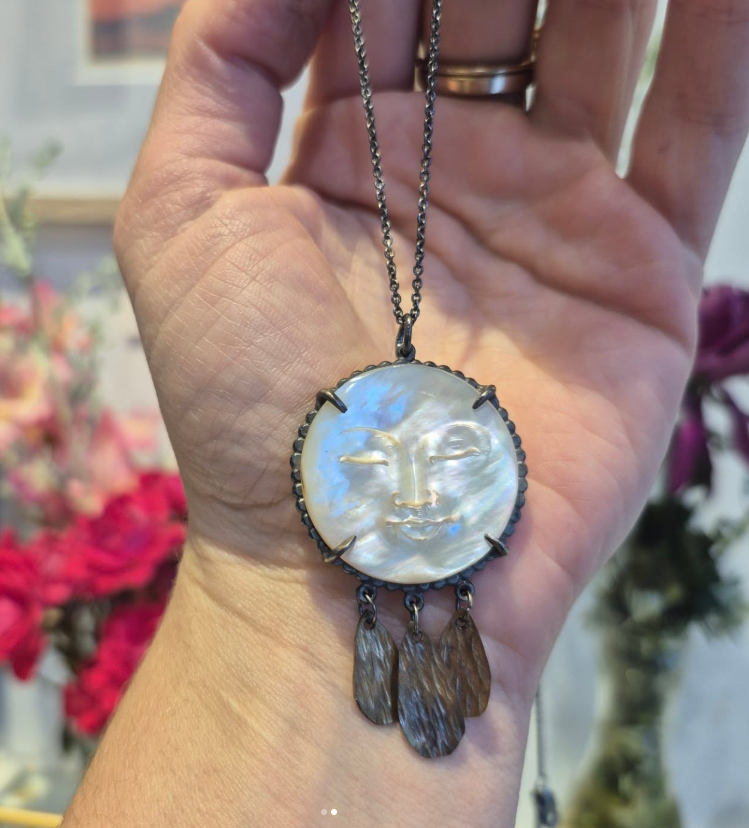 Hand holding a moon face necklace with a face pendant against a blurred floral background by Alice Scott