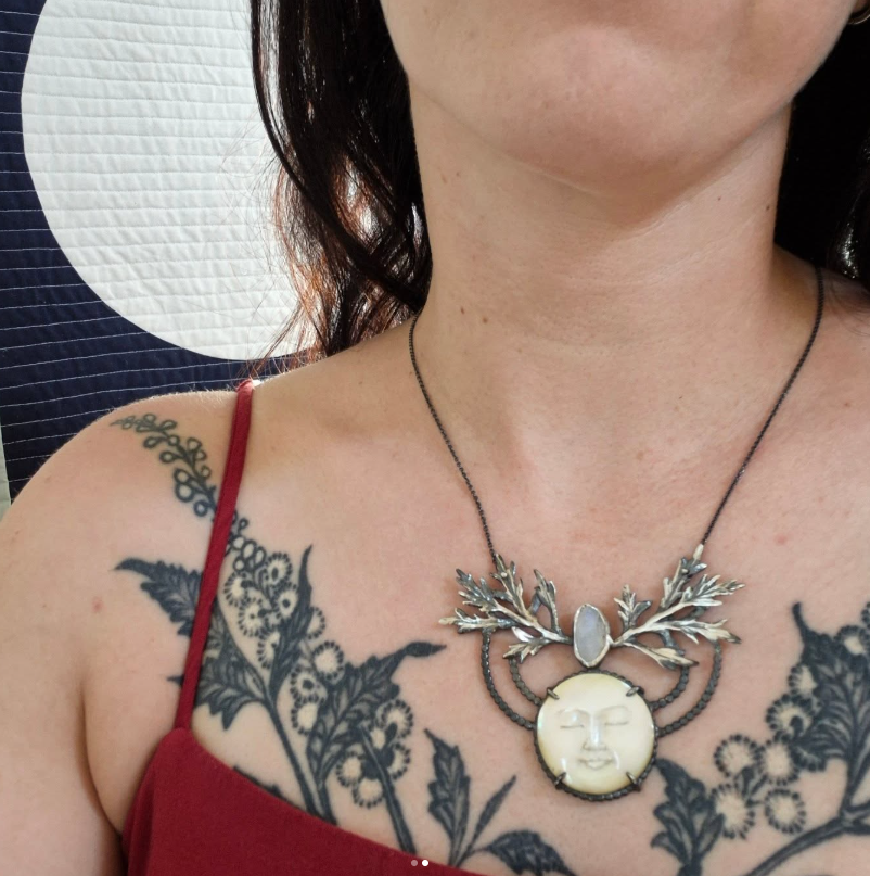 Person wearing a moon face necklace with a pendant featuring a face design, with tattoos on the shoulder and chest, by Alice Scott