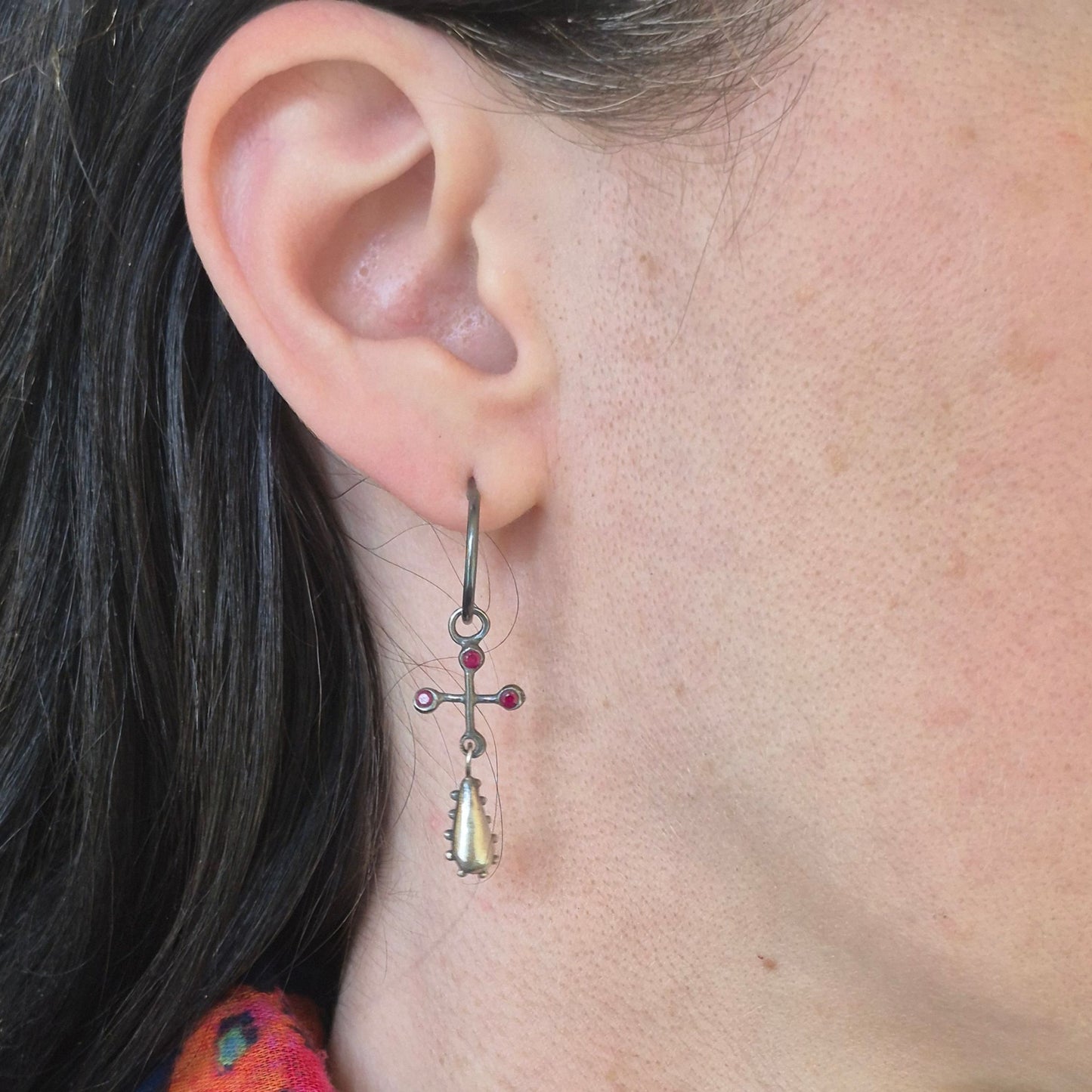 Close-up of an ear wearing a dangling earring with a cross design and rubies by Alice Scott