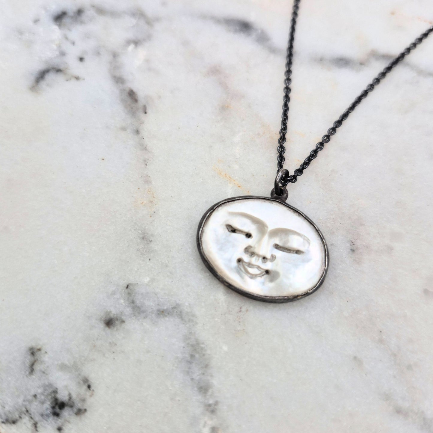 A necklace with a circular pendant featuring a carved peaceful face design on mother of pearl, with a black patina on sterling silver by Alice Scott