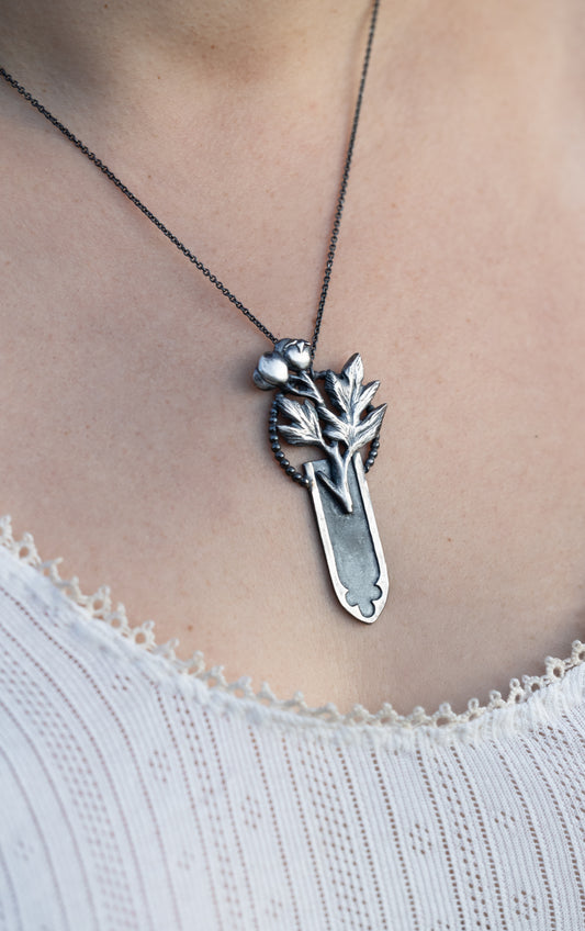 Silver necklace with a hawthorn leaf emerging from gothic arch, worn by a person wearing a white top., by Alice Scott