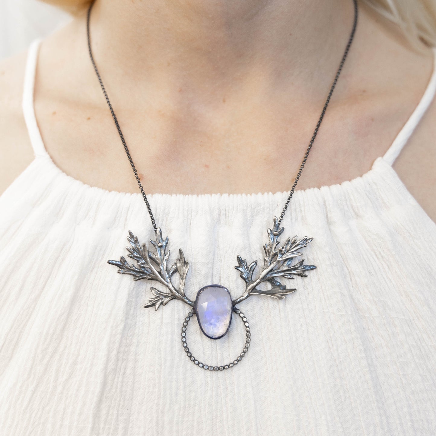 A person wearing a white dress showcasing a silver necklace with a mugwort design and a central moonstone with blue flash.