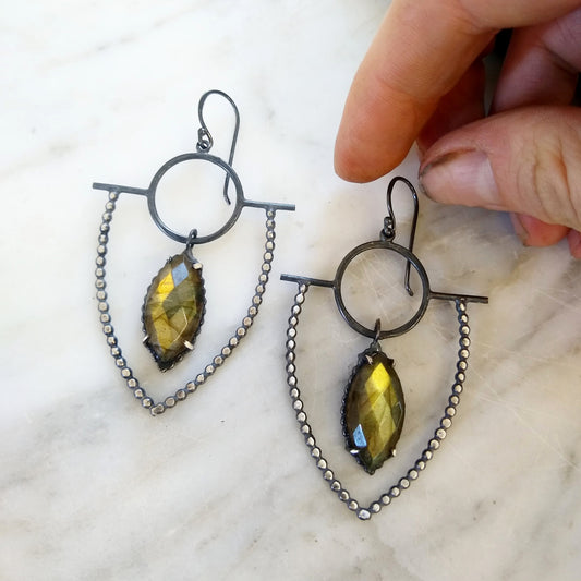 dotted blackened silver earrings that look like a shield with marquise green labradorite stones, on marble background with hand, by Alice Scott