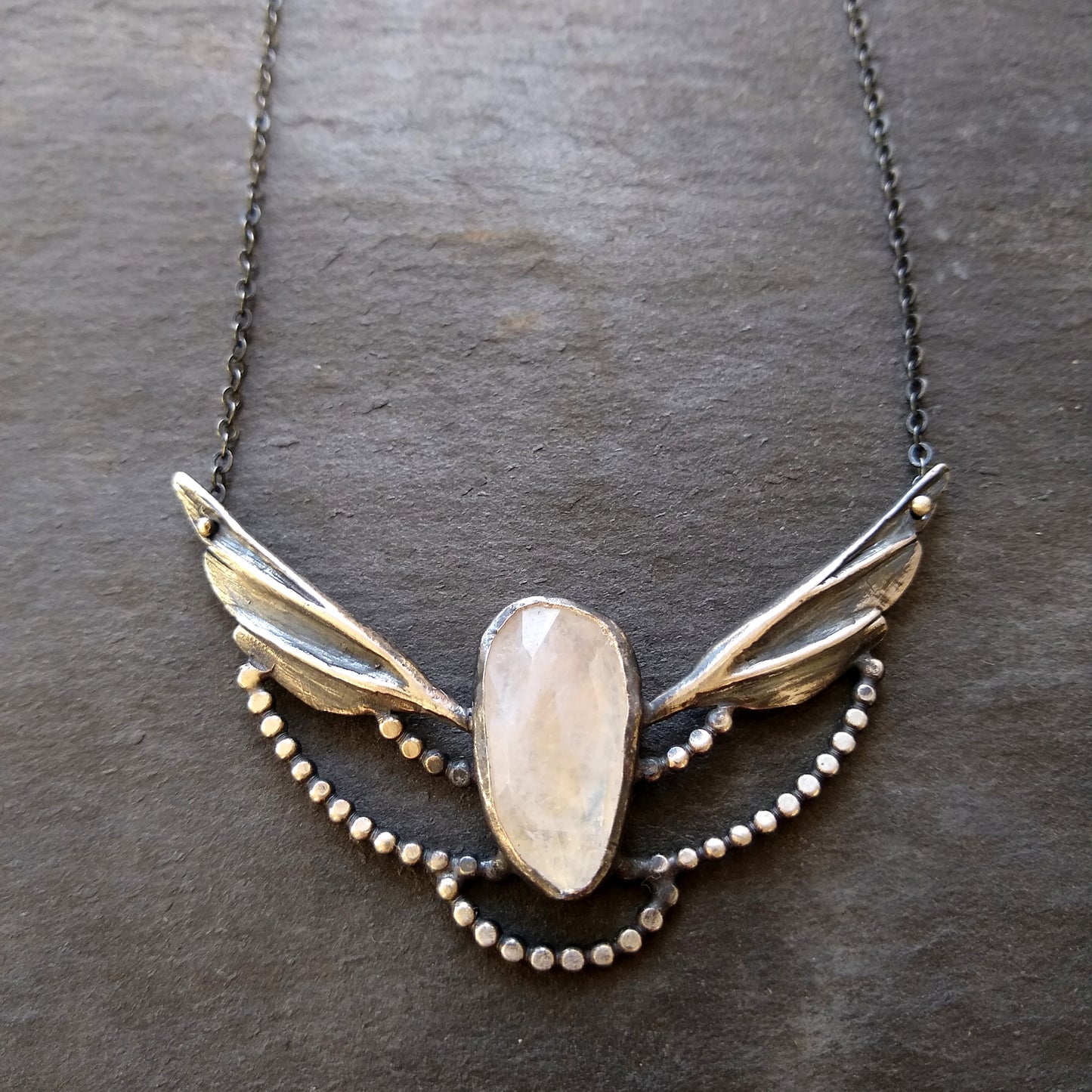 A silver necklace with a moonstone wing pendant and small silver accents on a slate background by Alice Scott