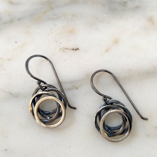 A pair of sterling silver earrings with a black patina, featuring a flowing circle design, displayed on a marble surface by Alice Scott