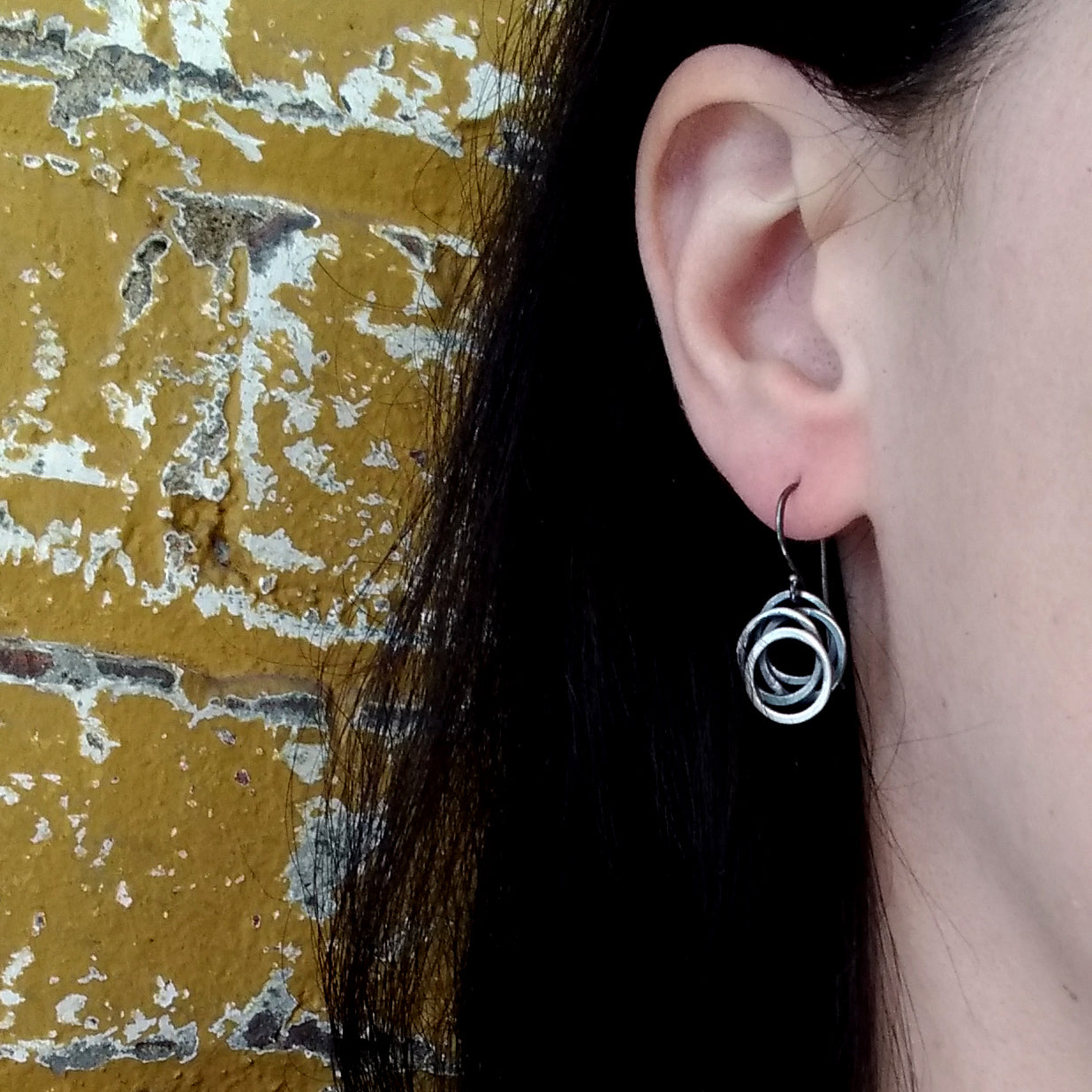 A pair of sterling silver earrings with a black patina, featuring a flowing circle design by Alice Scott, displayed on a model ear.