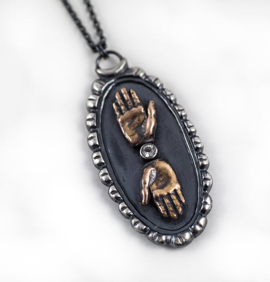 Close-up of a double hand cameo necklace with bronze hands and a sterling silver cameos background, featuring a topaz gemstone between the hands, by Alice Scott, on a white background