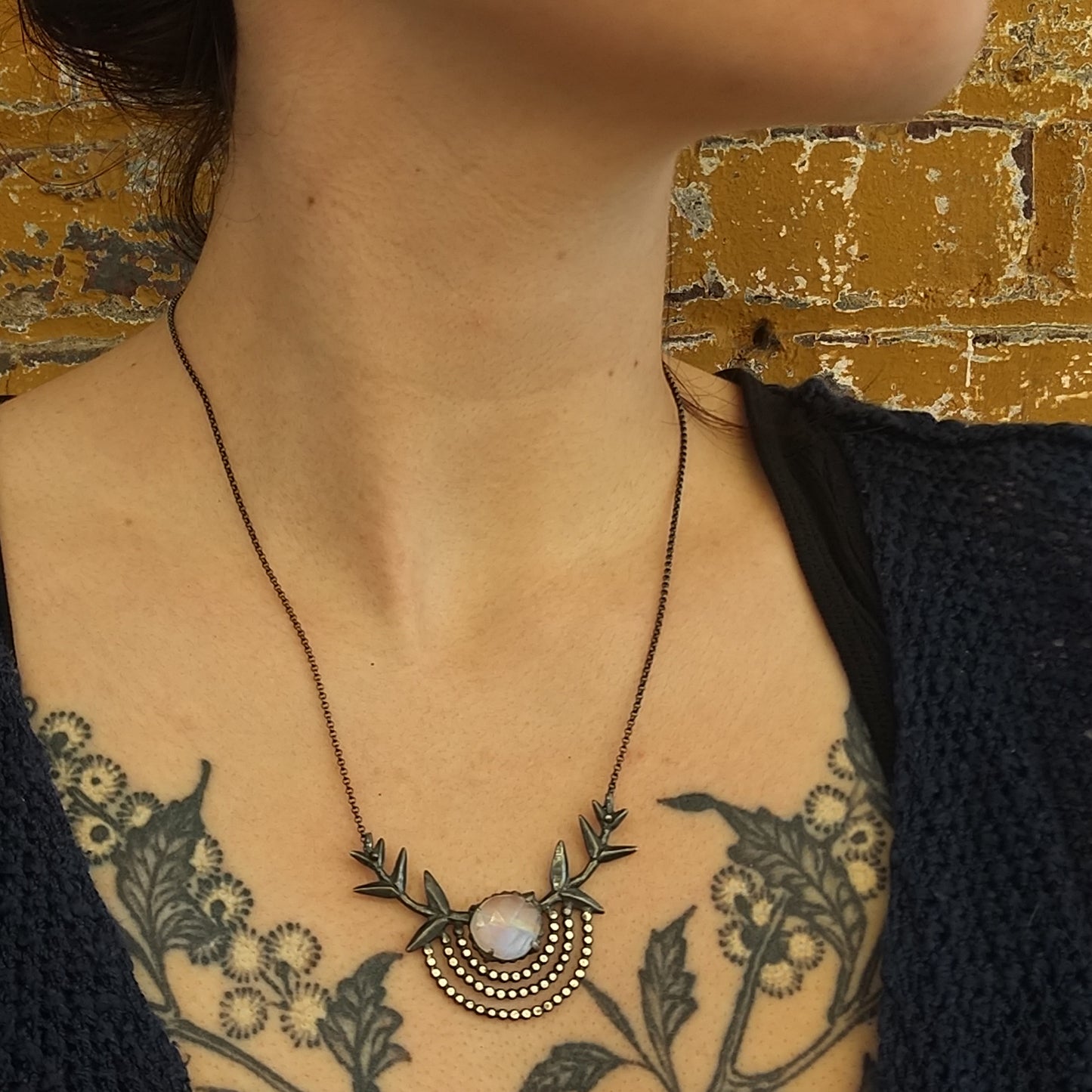 A silver necklace with a black patina featuring a branch pendant and a central moonstone gem, displayed against a model neck by Alice Scott
