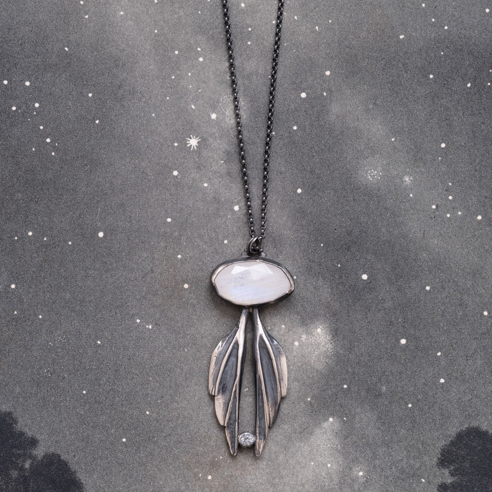 A sterling silver necklace with a moonstone pendant carved into the shape of wings, suspended from a chain against a starry background, by Alice Scott