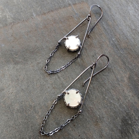 A pair of dangle earrings featuring mother of pearl circles connected to sterling silver chains, with a black patina finish, displayed against a textured grey background, by Alice Scott