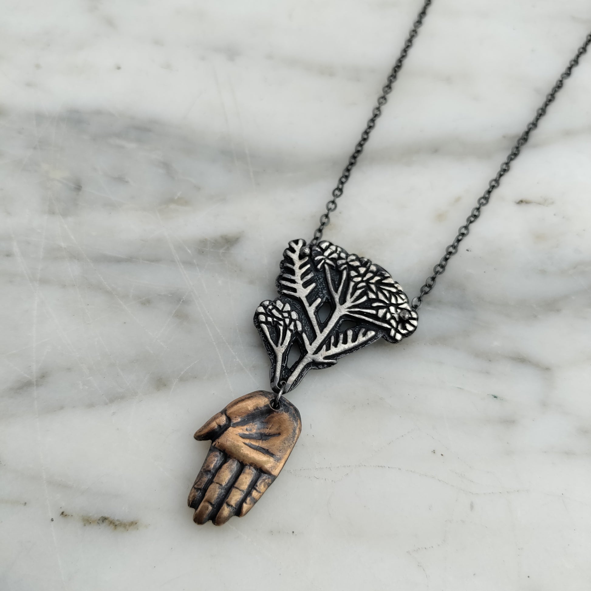 Bronze hand and silver yarrow pendant on marble background by Alice Scott.