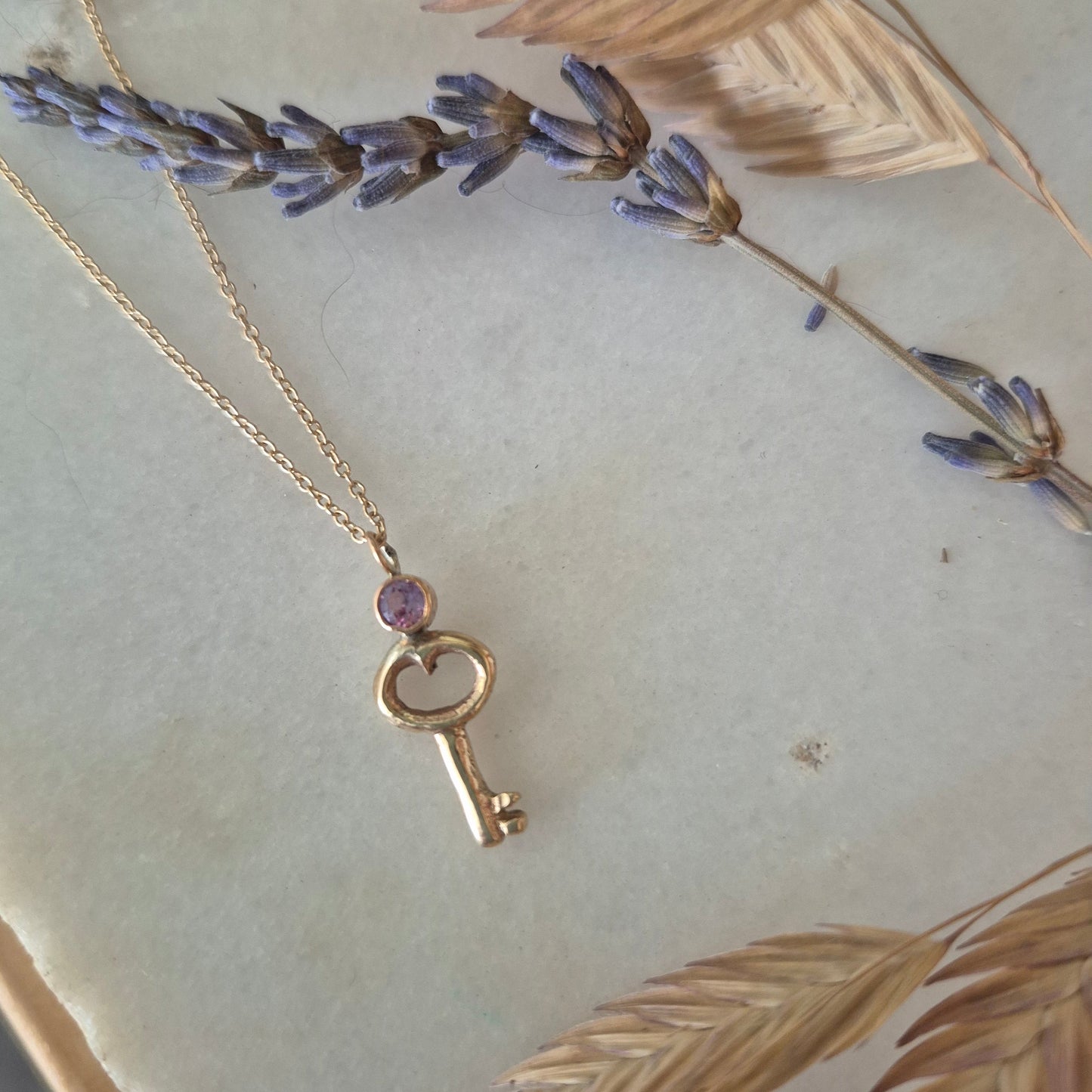 Gold key-shaped pendant with a purple gemstone on a chain, set against a decorative background with leaves and flowers.