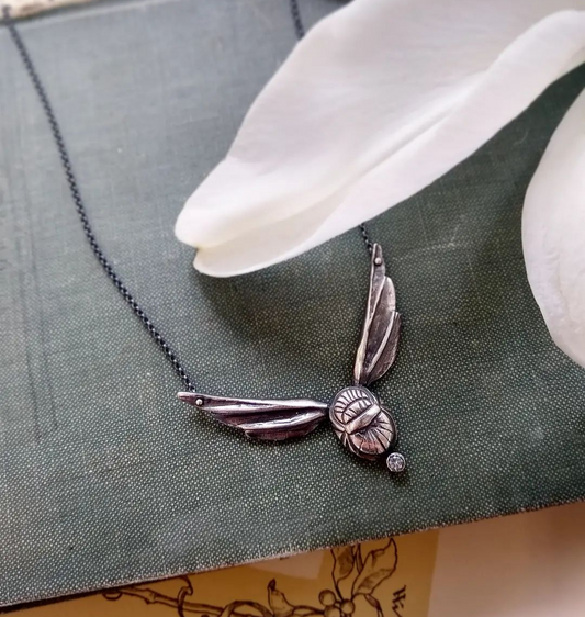 A sterling silver scarab wing necklace with a white topaz stone, displayed on a book by Alice Scott