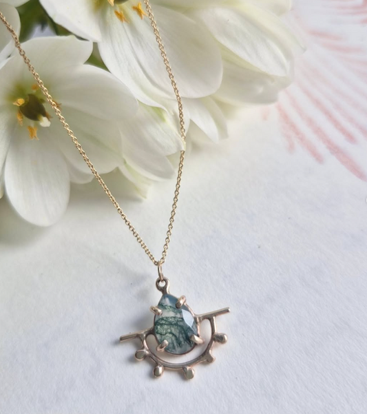 A gold necklace with a pendant featuring a moss agate stone, presented on a card with white flowers in the background by Alice Scott