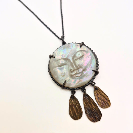 Necklace with a round mother of pearl pendant featuring a moon face design and brass droplets on a white background by Alice Scott