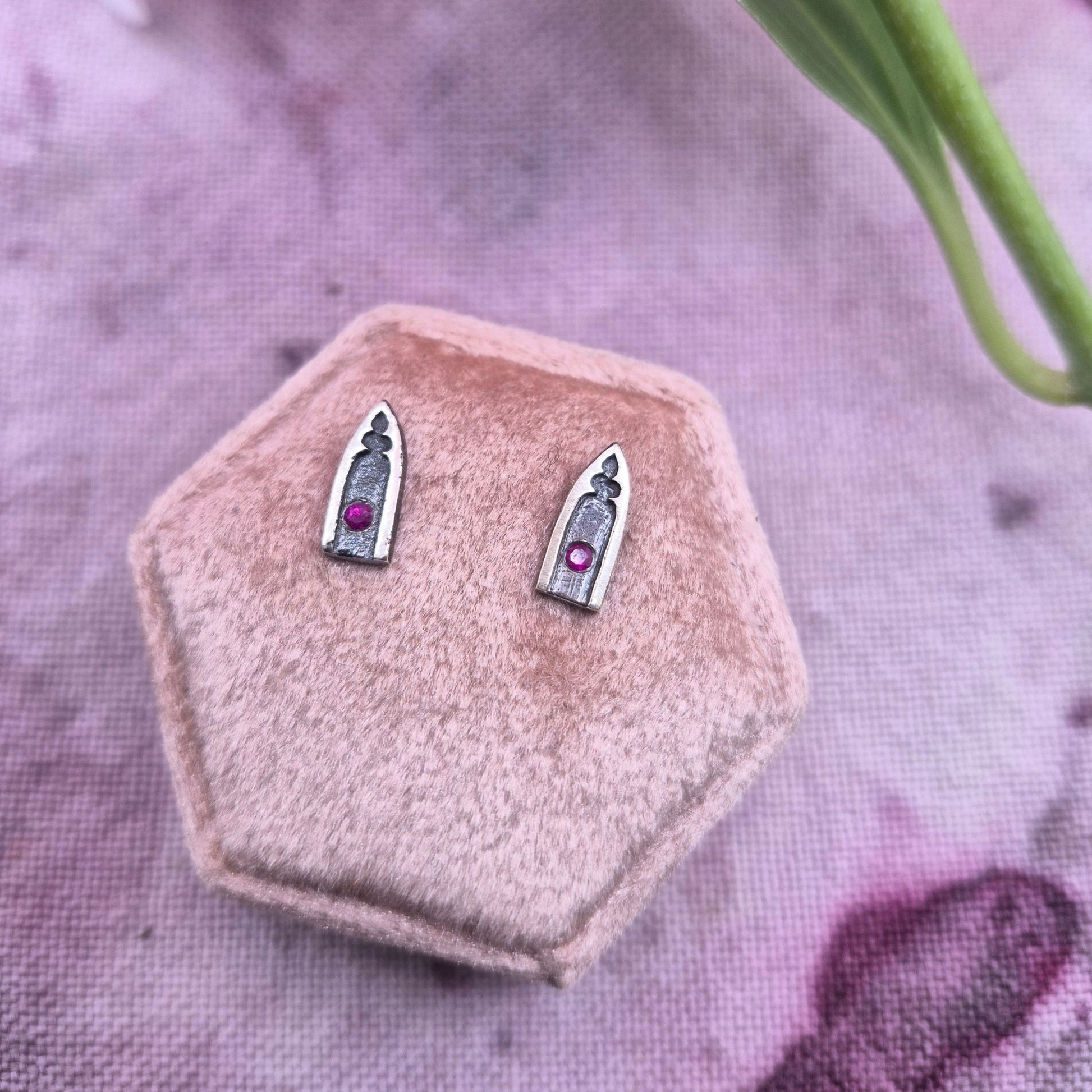 Silver gothic arch earrings with red ruby gemstones on a pink hexagonal stand against a purple fabric background by Alice Scott