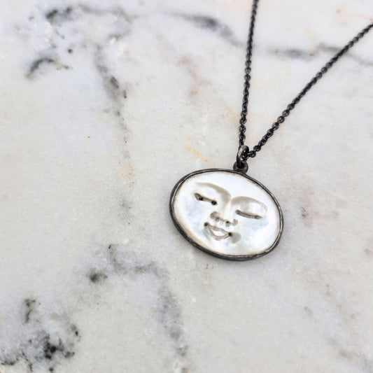 A necklace with a circular pendant featuring a carved peaceful face design on mother of pearl, with a black patina on sterling silver by Alice Scott