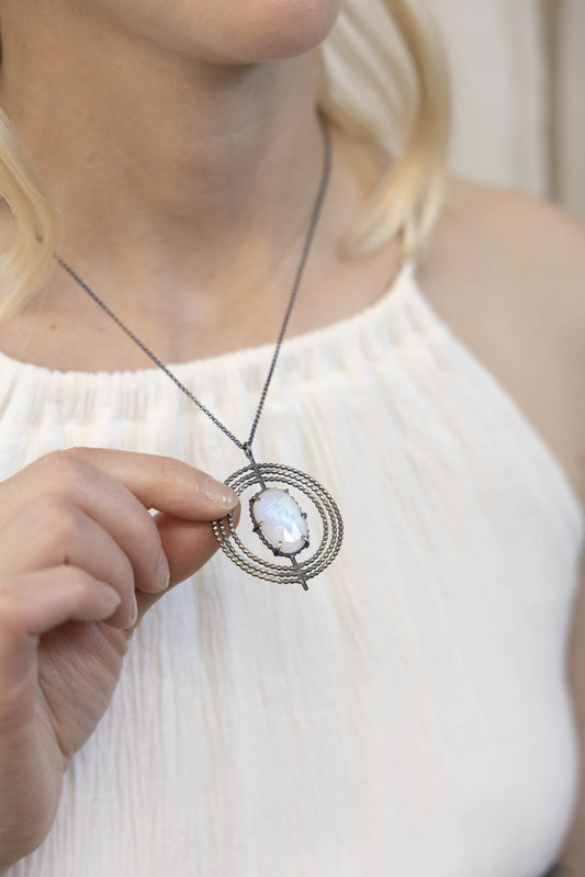 A necklace with a central moonstone surrounded by a circular silver frame with intricate details, on a model,  by Alice Scott