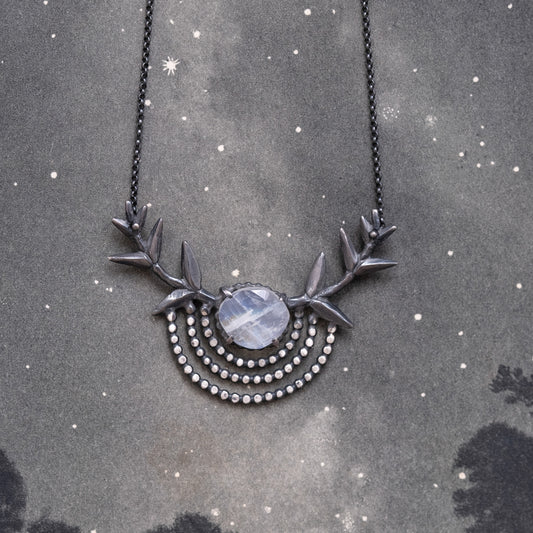 A silver necklace with a black patina featuring a branch pendant and a central moonstone gem, displayed against a dark textured background with stars by Alice Scott