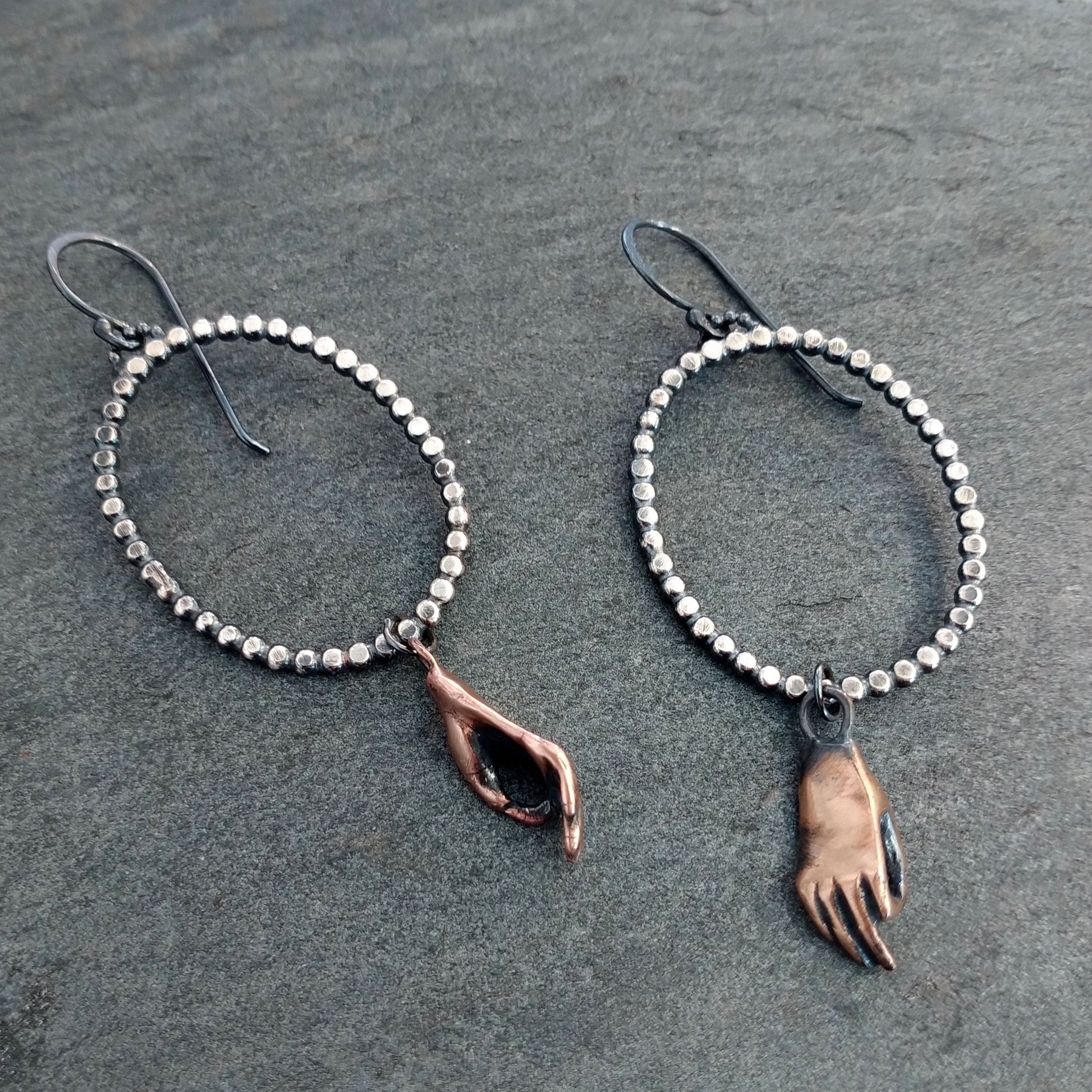 Dotted silver oval earrings with bronze cast meditating hands hanging, by Alice Scott on textured gray background.
