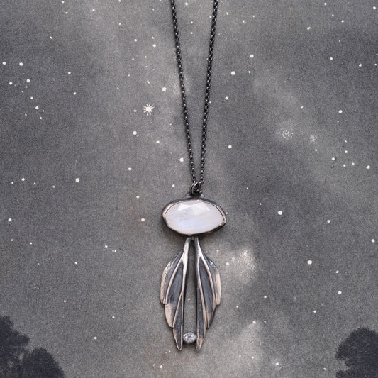 A sterling silver necklace with a moonstone pendant carved into the shape of wings, suspended from a chain against a starry background, by Alice Scott